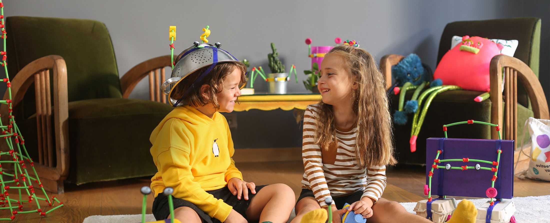 Two children laughing on a living room rug, one wearing a colander helmet, surrounded by toys they built with Toyi